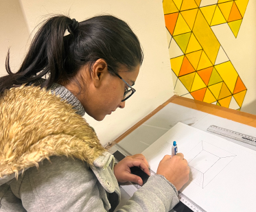 A person in a gray hoodie draws a geometric shape at a desk, with a yellow and orange triangular pattern on the wall.