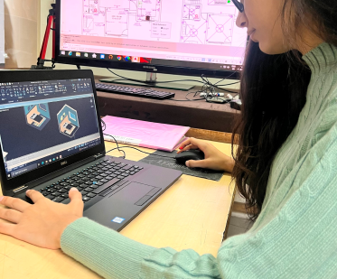 A person in a light green sweater uses CAD software on a laptop, with a technical drawing visible on a monitor in the background.