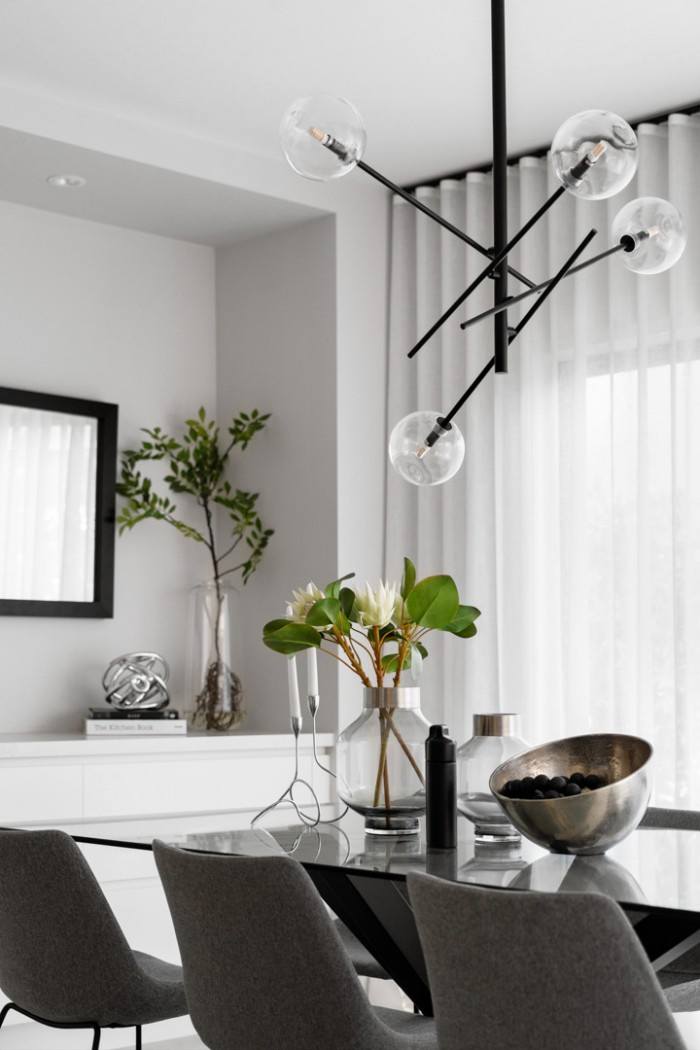 A modern dining room features a glass table with gray chairs, a metallic fruit bowl, a vase with green leaves and white flowers, and a geometric chandelier.