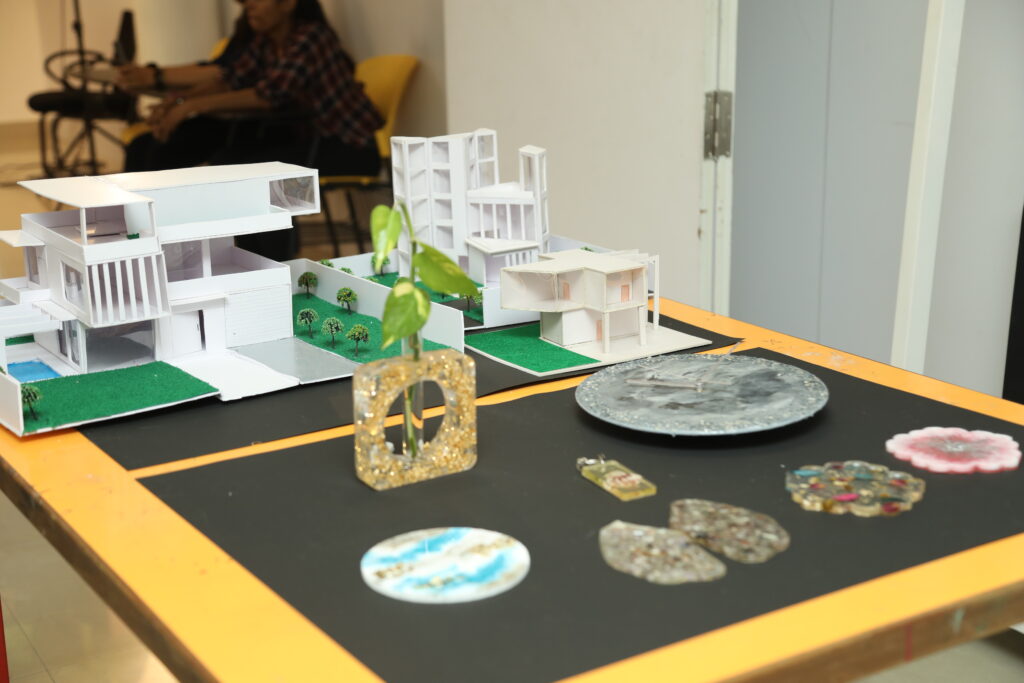 A table displays modern building models, jewelry, and resin art on a black mat. A partially visible person sits with a door ajar.