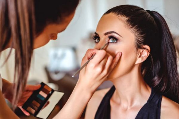 Close-up of a woman with tied-back hair getting makeup applied with a thin brush, as the artist holds an eyeshadow palette.