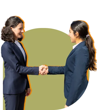 Two women in business attire shake hands, standing opposite each other, and smiling.