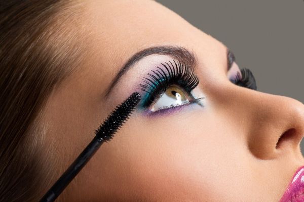 Close-up of a woman applying mascara, with blue and purple eye makeup, pink lips, and brown eyes looking up, against a gray background.