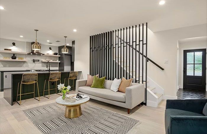 Modern open-plan: green kitchen island, gray sofa, patterned rug, black staircase, dark green chair, pendant lights.