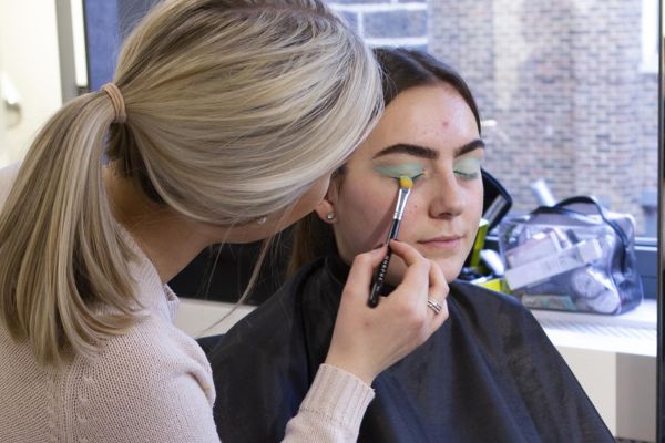 A blonde applies green eyeshadow to another person in a black cape, with makeup tools and a window in the background.