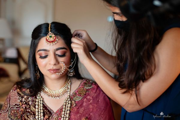 A bride in traditional attire with intricate jewelry has her headpiece adjusted, eyes closed, looking serene.