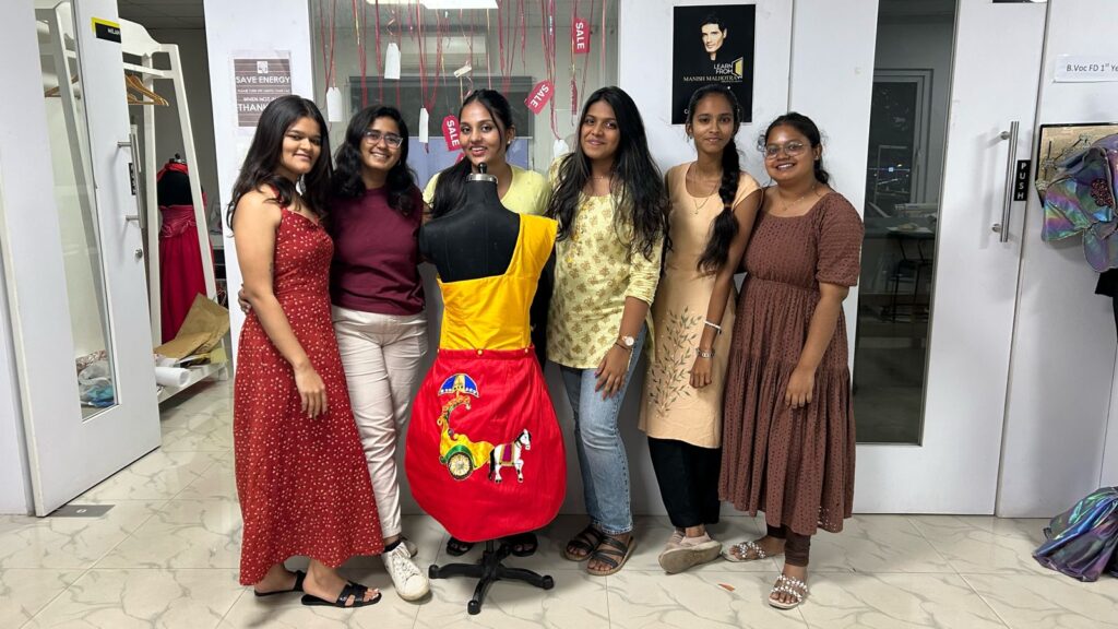 Six women smile around a mannequin in a vivid dress, surrounded by garments in a fashion studio.