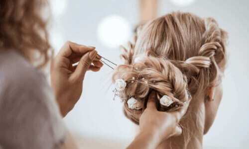 A stylist secures a blonde woman's updo with white flowers using a hairpin, with a softly blurred background.