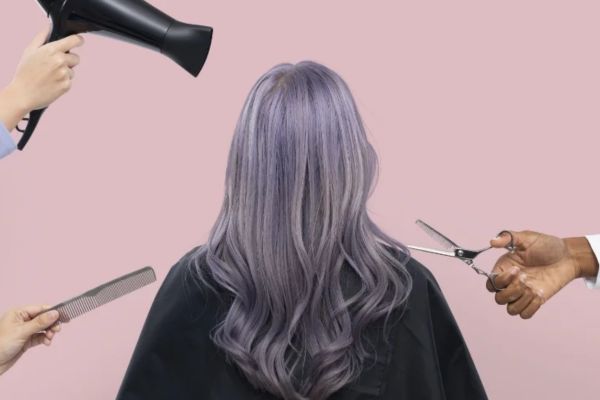 Person with long pastel purple hair sits with a pink background. Hands with a hairdryer, comb, and scissors indicate a styling session.