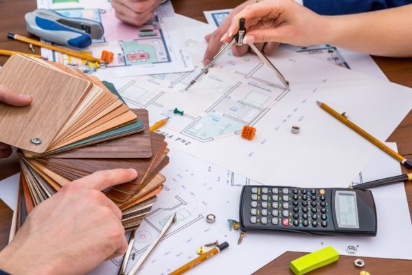 A cluttered table with floor plans, a calculator, pencils, a compass, a stapler, a protractor, colored swatches, and hands working.