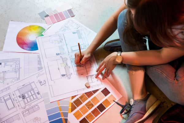 A person in jeans and a watch sits on the floor, drawing plans with color swatches, design tools, and samples nearby.