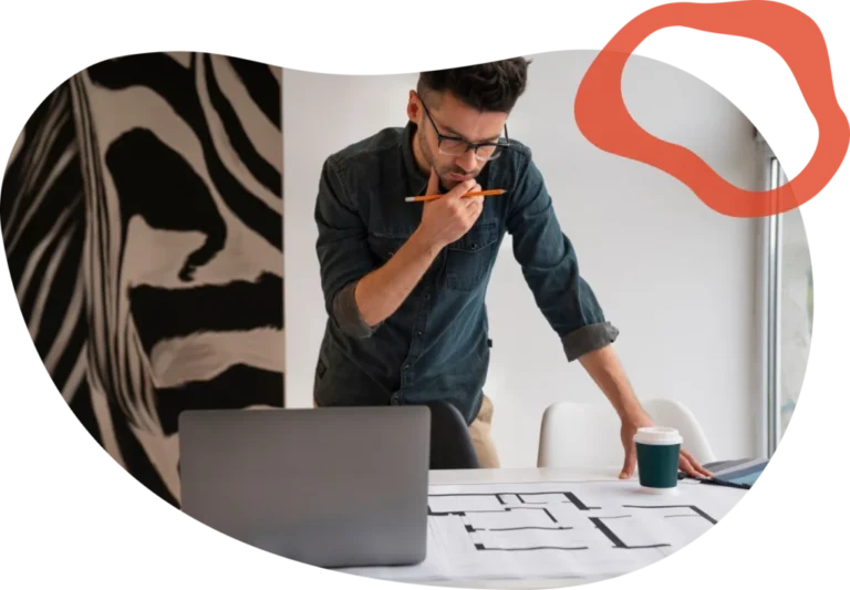 A man in glasses and a denim shirt studies plans at a desk, holding a pencil, with a laptop, coffee cup, and zebra-patterned wall in view.