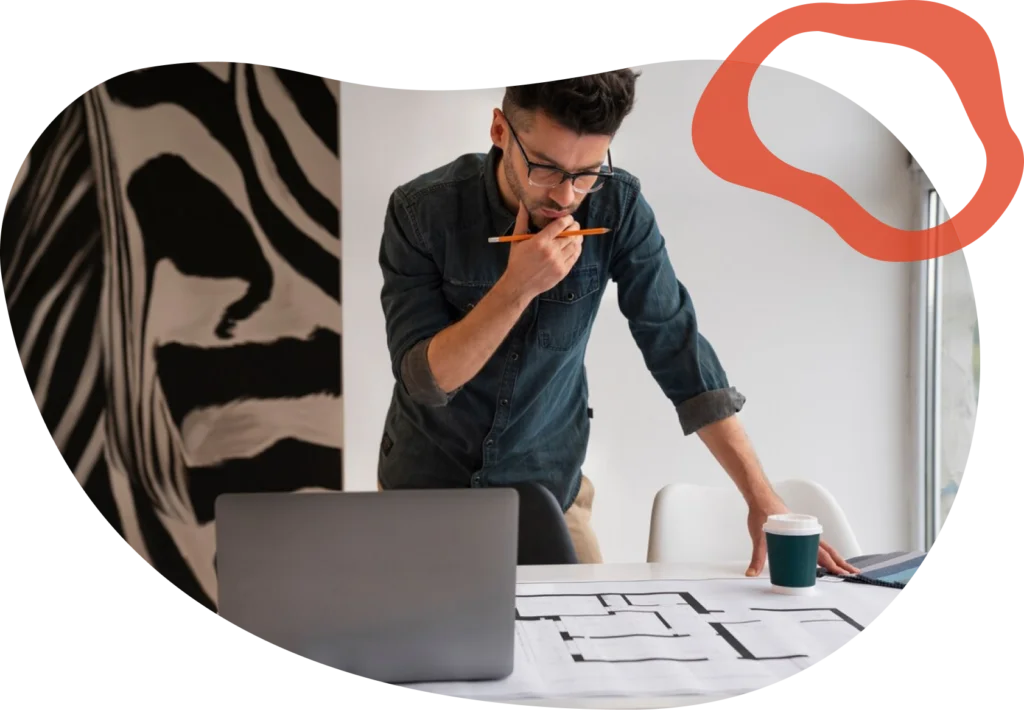 A man in glasses and a denim shirt studies plans at a desk, holding a pencil, with a laptop, coffee cup, and zebra-patterned wall in view.